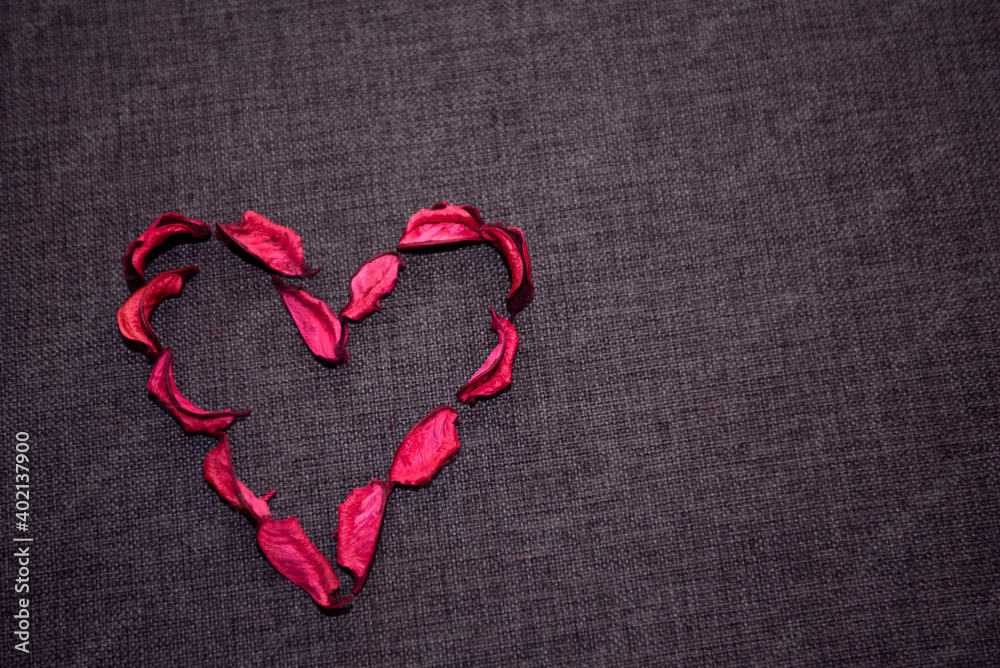 Символ сердца  из сухих  лепестков красной  розы на сером текстильном  фоне.