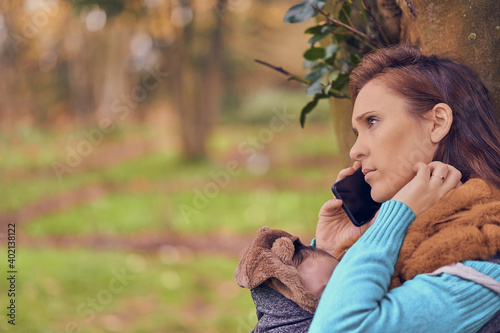 beautiful woman talking on the smart phone in winter in a park wearing coat and pullover. carrying her child. maternity lifestyle © Patricia