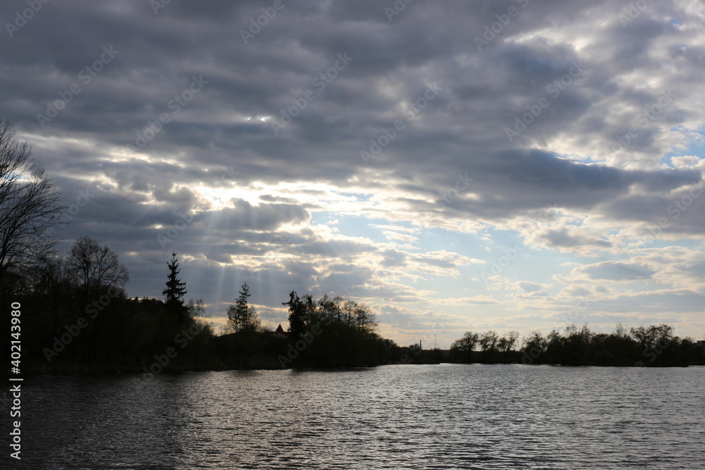 
The sun's rays make their way through the clouds in the sky on a spring evening on the shore of a reservoir