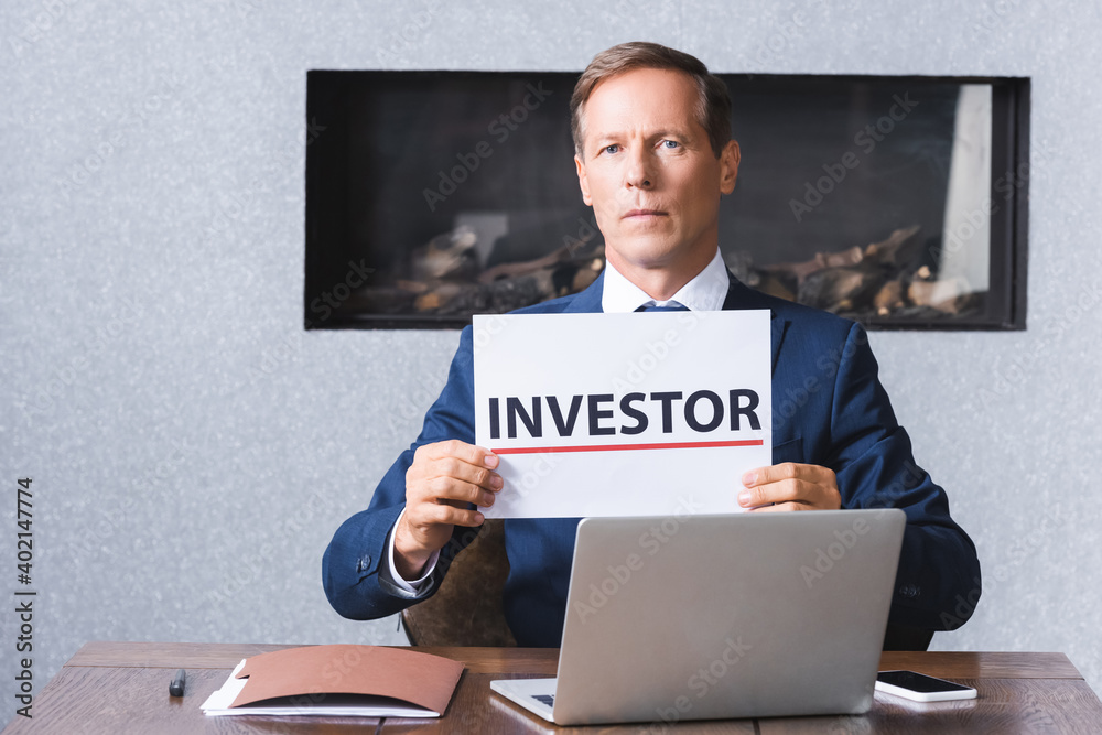 Serious businessman looking at camera and holding sign with investor lettering at workplace with digital devices in meeting room. Serious businessman looking at camera and holding sign with investor lettering at workplace with digital devices in meeting room.