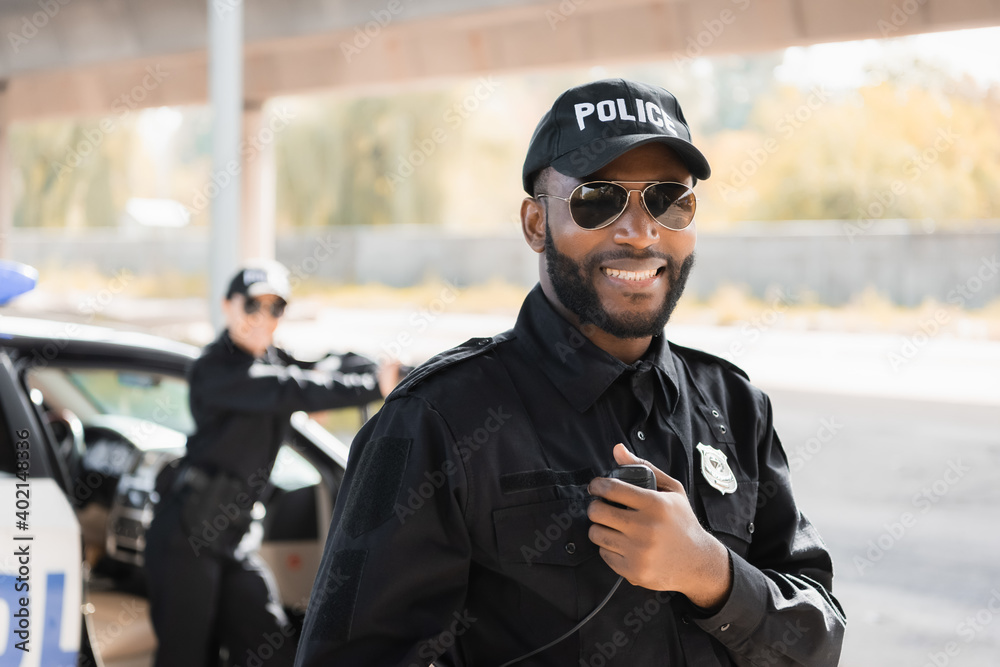 happy african american police officer looking at camera while holding radio  set on blurred background outdoors. Stock Photo | Adobe Stock