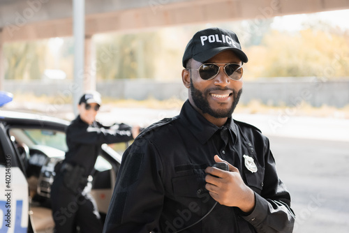 Print op canvas happy african american police officer looking at camera while holding radio set on blurred background outdoors
