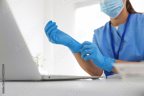 Doctor in protective mask putting on medical gloves at table in office, closeup