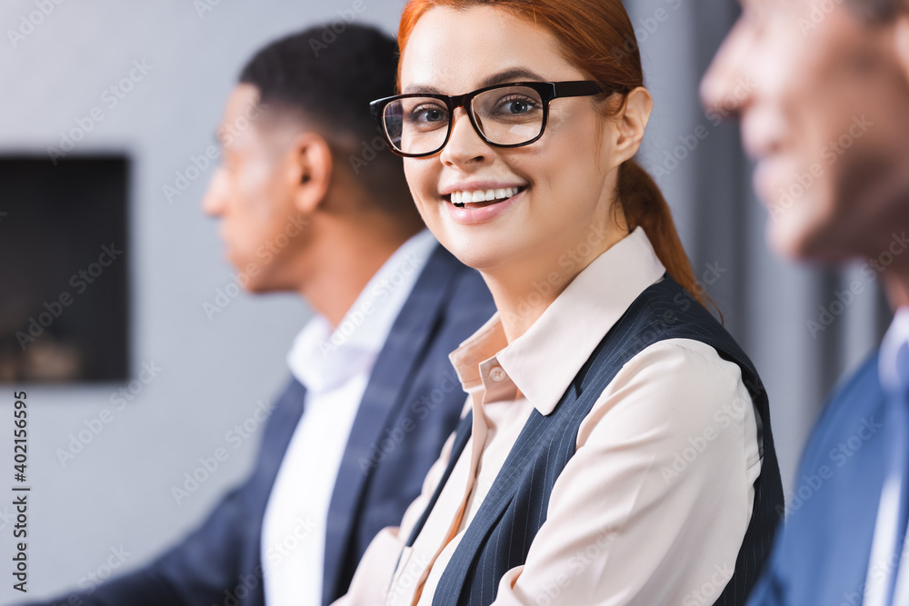 Smiling redhead businesswoman in eyeglasses looking at camera with blurred african american colleague on background. Smiling redhead businesswoman in eyeglasses looking at camera with blurred african american colleague on background.