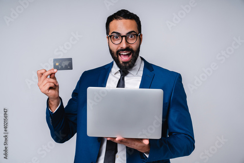 Overexcited young handsome Arab businessman holding credit card and laptop isolated over grey background photo