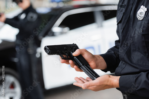 Cropped view of policeman loading gun near colleague and police car on blurred background.