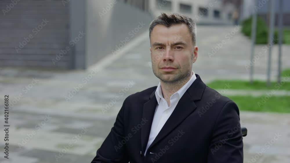 Portrait of serious guy in formal suit turning head and looking to camera while sitting in wheel chair. Crop view of disabled businessman near office building. Zoom in. Portrait of serious guy in formal suit turning head and looking to camera while sitting in wheel chair. Crop view of disabled businessman near office building. Zoom in.