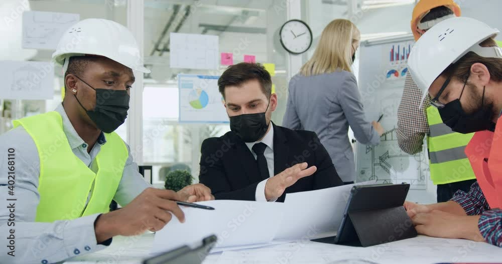 Work during coronavirus pandemia where good-looking confident hardworking multiethnic engineers and their main manager or ceo in protective masks brainstorming about building project during meeting