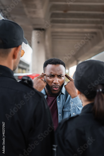 Carta da parati angry african american victim showing you are insane gesture while arguing with police officers on blurred foreground outdoors
