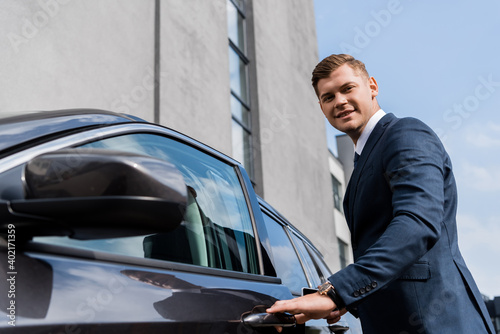 smiling businessman looking at camera while opening car on blurred foreground. © LIGHTFIELD STUDIOS