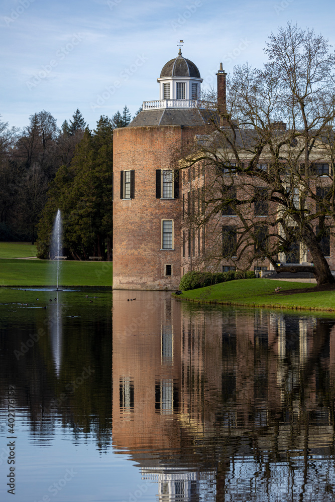 Vertical frame with mirroring reflection of the Rosendael castle behind barren winter trees in the still water of the garden pond in the foreground against a blue sky