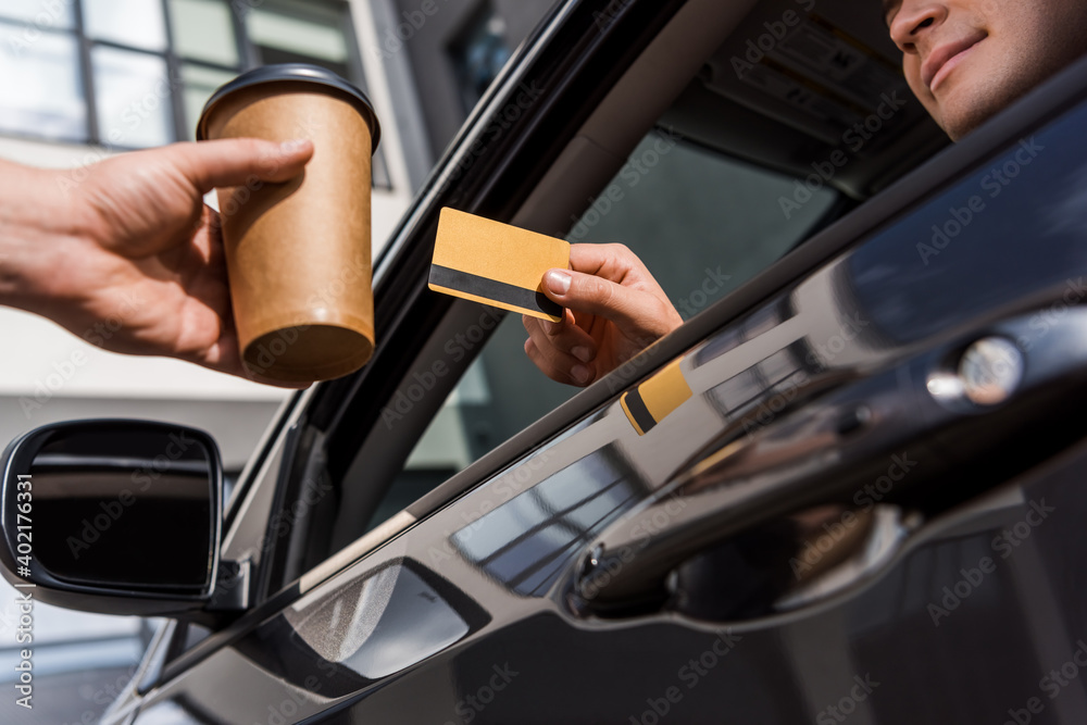 Cropped view of man holding credit card while sitting in car near seller with coffee to go on blurred foreground.