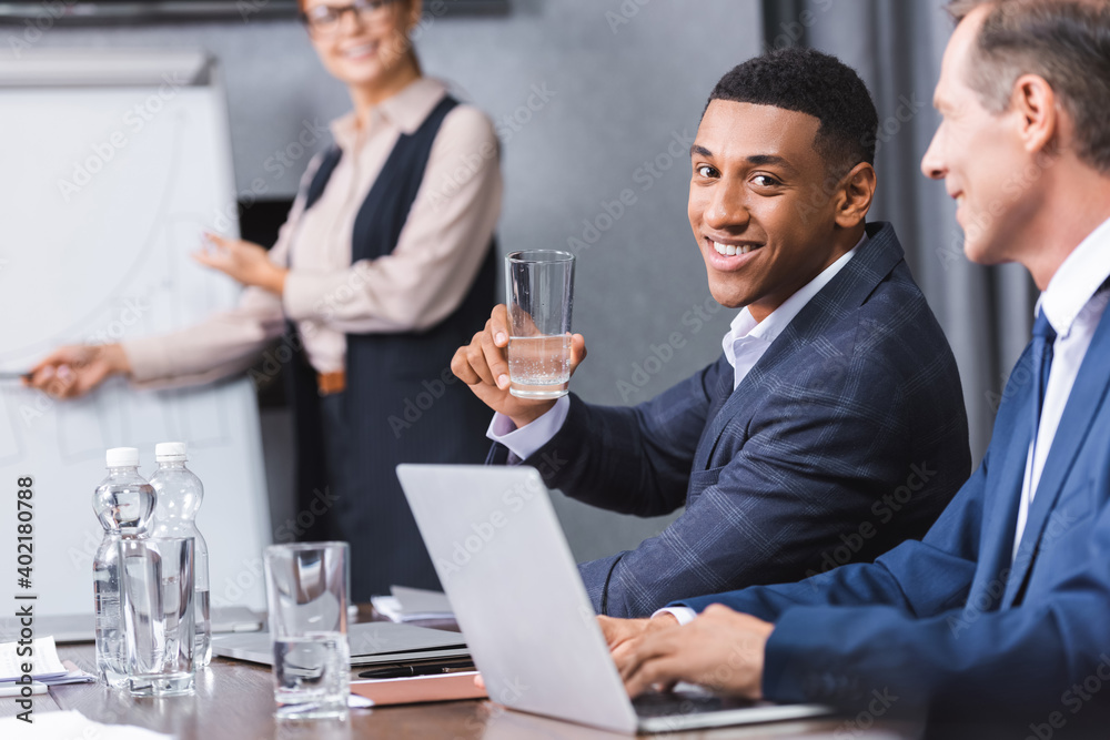 Smiling african american businessman with glass of water looking at camera while sitting at workplace on blurred background. Smiling african american businessman with glass of water looking at camera while sitting at workplace on blurred background.