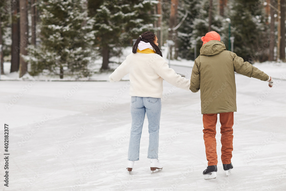 Rear view of couple in warm clothing skating together on skating rink in winter