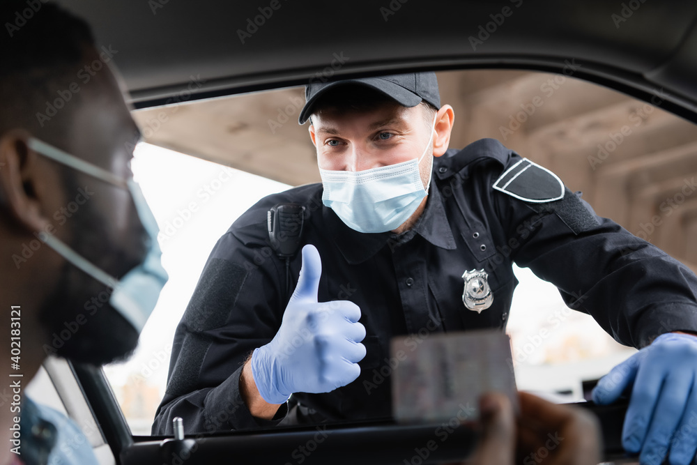 Police officer in medical mask and latex gloves showing like to african american driver holding license on blurred foreground in car. Police officer in medical mask and latex gloves showing like to african american driver holding license on blurred foreground in car.