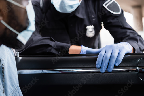 Cropped view of policeman in latex gloves standing near african american driver in medical mask on blurred foreground in car.