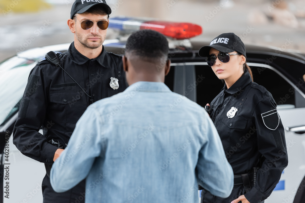 back view of african american victim standing near serious police officers on blurred foreground outdoors.