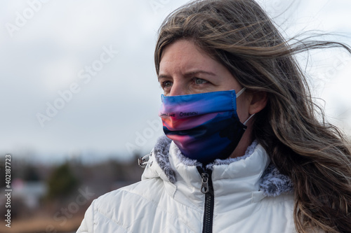 A close up head shot of a woman with long hair wearing a colorful protective face mask and a white winter coat