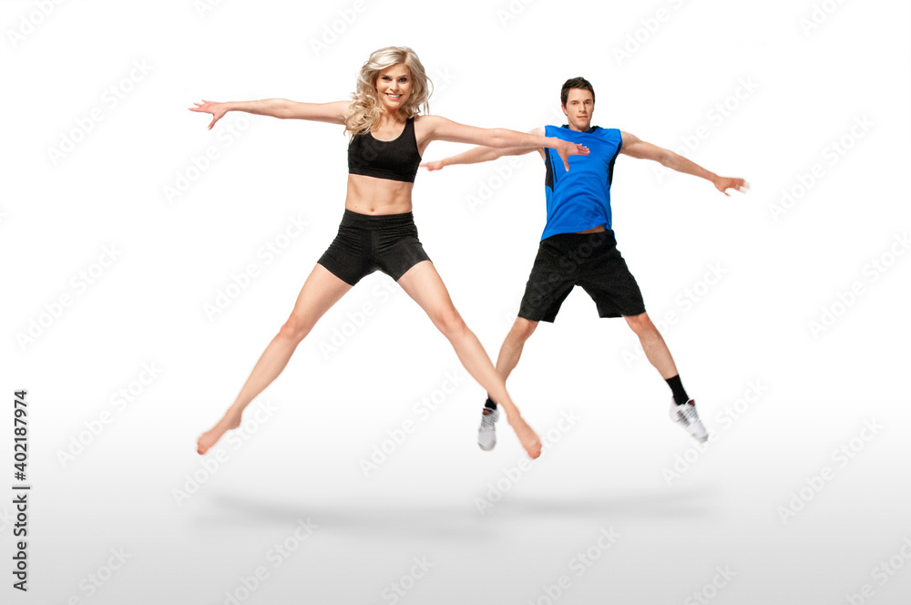 Portrait of a fit, young, white coach in a blue shirt & female athlete with curly long blond hair posing together jumping in a studio with a white background wearing black shorts & sports bra.