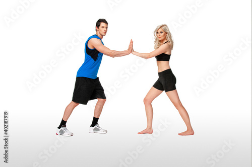 Portrait of a fit, young, white coach in a blue shirt & female athlete with curly long blond hair posing together touching palms in a studio with a white background wearing black shorts & sports bra.