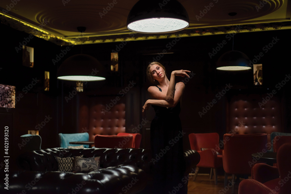 A graceful ballerina in a black dress posing in a restaurant. Beautiful elegant woman dancing in a dark room