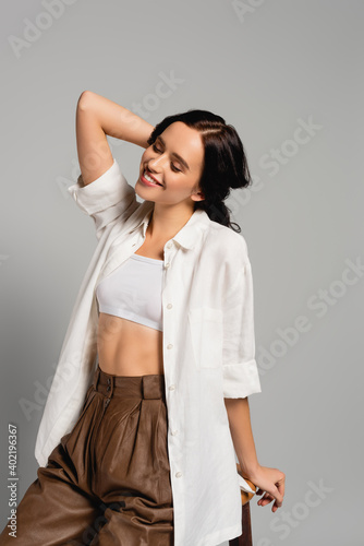  brunette woman with closed eyes wearing casual clothes and posing while leaning on stool isolated on grey