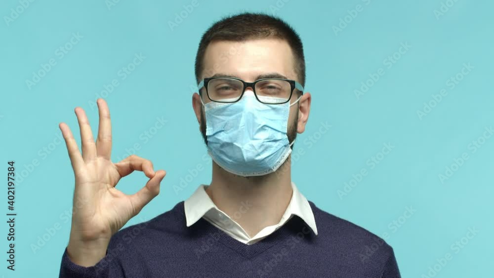 Covid-19, quarantine and pandemic concept. Slow motion closeup of handsome man in glasses and medical mask standing confident, showing okay sign, guarantee quality, blue background Covid-19, quarantine and pandemic concept. Slow motion closeup of handsome man in glasses and medical mask standing confident, showing okay sign, guarantee quality, blue background