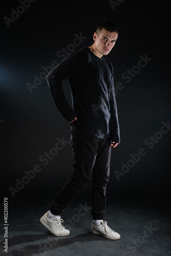 Full-length picture of an attractive sexy young male model posing isolated on a black background and wearing black blouse and black pants.