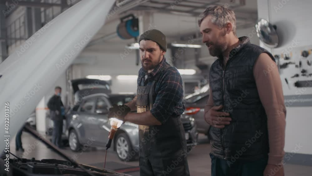 Handheld shot of male auto mechanic in overalls and gloves holding portable work light and looking under hood of car while talking to client in garage Handheld shot of male auto mechanic in overalls and gloves holding portable work light and looking under hood of car while talking to client in garage