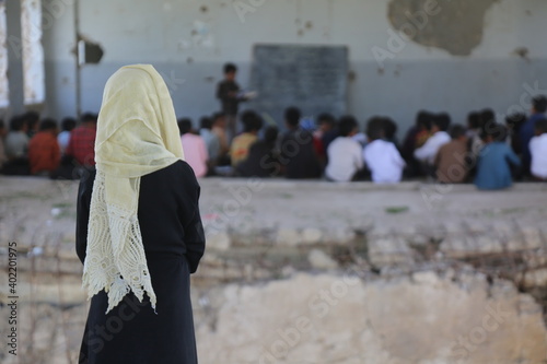  A sad Yemeni child sits on the rubble of her destroyed school and watches her friends study there