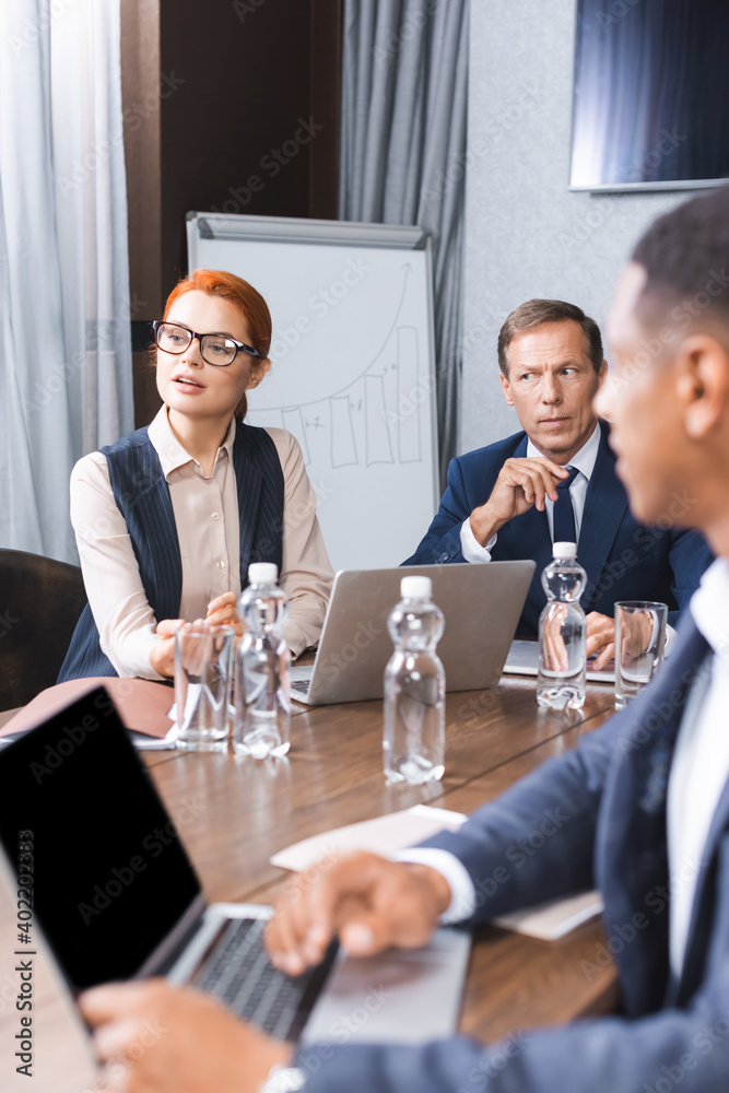 Redhead businesswoman talking while sitting at workplace with multicultural colleagues on blurred foreground in meeting room. Redhead businesswoman talking while sitting at workplace with multicultural colleagues on blurred foreground in meeting room.