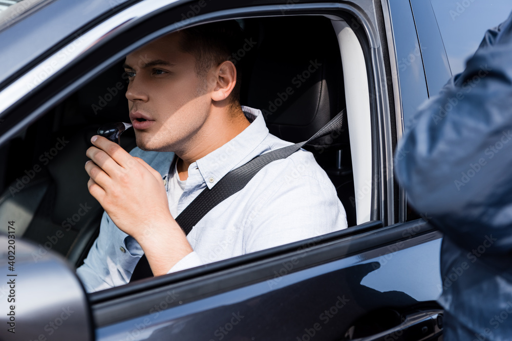 young man in car blowing into breathalyzer, and policeman standing on blurred foreground.