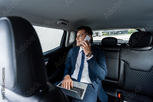 Smiling businessman talking on smartphone and using laptop in car. © LIGHTFIELD STUDIOS