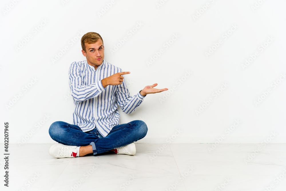 Young caucasian man sitting on the floor excited holding a copy space on palm.