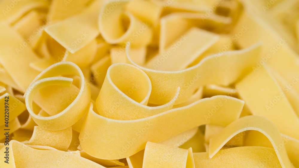 Detail of wide flat noodles pile in beautiful pasta background. Close-up of yellow curled wheat dough strips in abstract culinary texture. Raw dried ribbon tagliatelles. Healthy diet. Selective focus. Detail of wide flat noodles pile in beautiful pasta background. Close-up of yellow curled wheat dough strips in abstract culinary texture. Raw dried ribbon tagliatelles. Healthy diet. Selective focus.