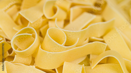 Detail of wide flat noodles pile in beautiful pasta background. Close-up of yellow curled wheat dough strips in abstract culinary texture. Raw dried ribbon tagliatelles. Healthy diet. Selective focus.