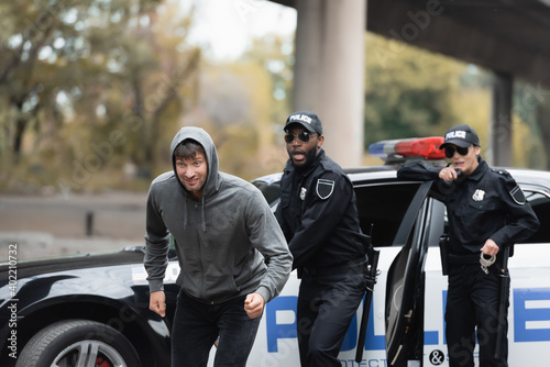 Φωτογραφία hooded offender running from shocked multicultural police officers on blurred background on urban street