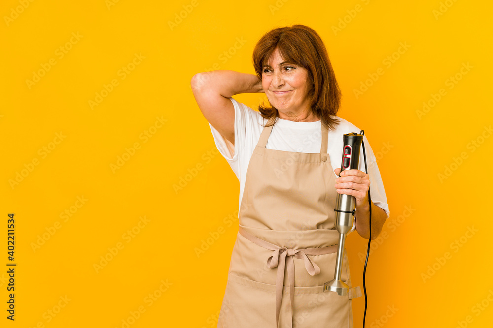 Middle age caucasian cook woman holding a mixer isolated touching back of head, thinking and making a choice. Middle age caucasian cook woman holding a mixer isolated touching back of head, thinking and making a choice.