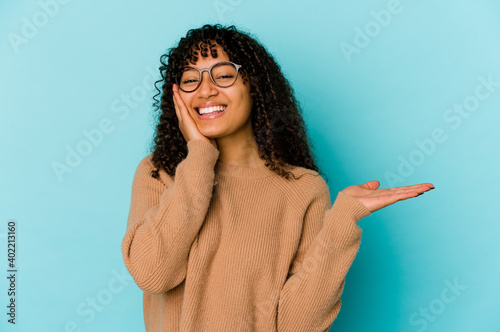 Young african american afro woman isolated holds copy space on a palm, keep hand over cheek. Amazed and delighted.