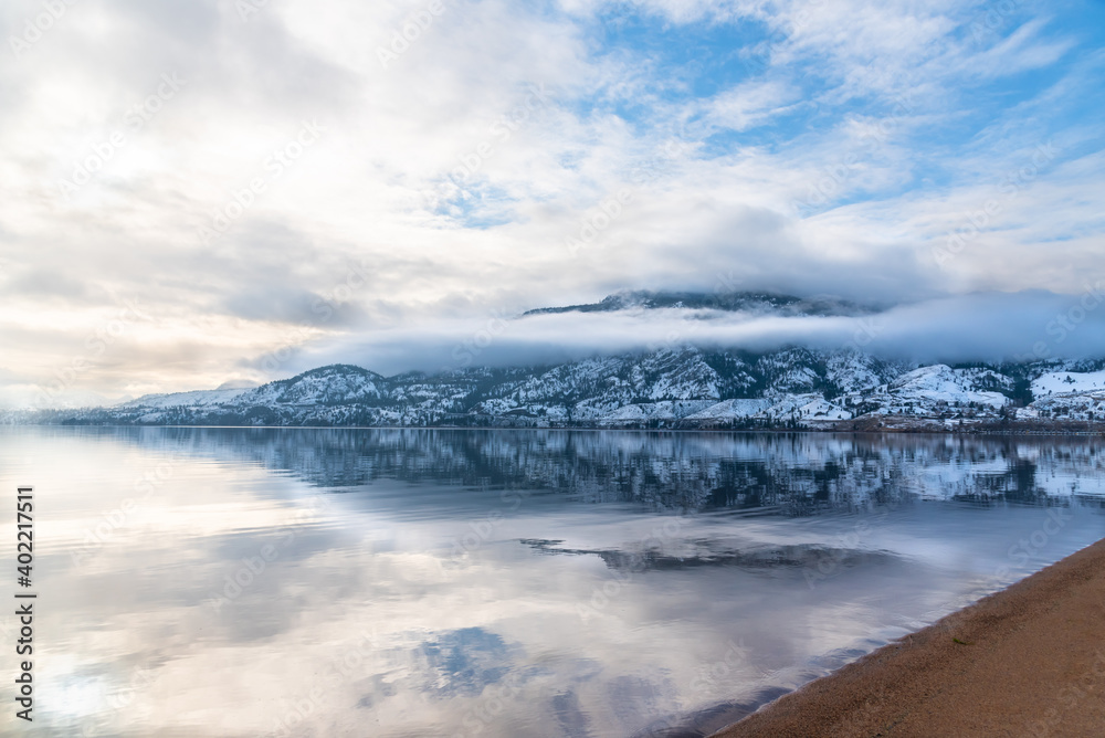 Snow covered mountain, fog, clouds, and sunlight reflected in calm lake