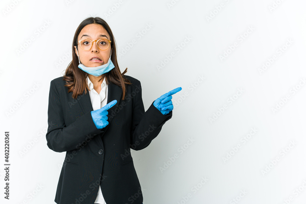 Young business latin woman wearing a mask to protect from covid isolated on white background shocked pointing with index fingers to a copy space. Young business latin woman wearing a mask to protect from covid isolated on white background shocked pointing with index fingers to a copy space.