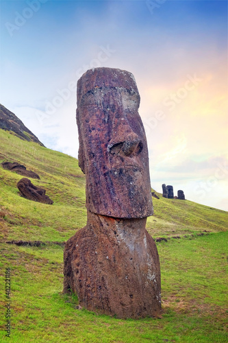 Moai on the slopes of the Rano Raraku Volcano, on Easer Island, surrounded by green vegetation, against a colorful sky.