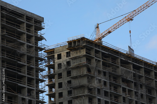 Two new concrete multi-storey buildings under construction with a crane lifting the construction plate.Gray high-rise structure with windows and ceilings in the working stage against the blue sky © Nina