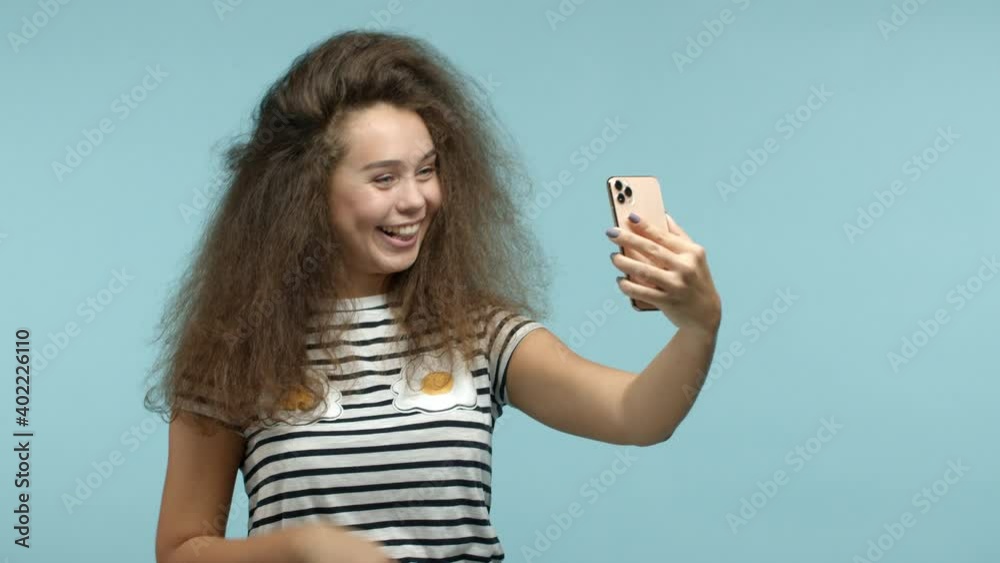 Slow motion of silly glamour girl talking to friends on video chat, waving hand at smartphone and saying hi, smiling joyfully, standing over blue background Slow motion of silly glamour girl talking to friends on video chat, waving hand at smartphone and saying hi, smiling joyfully, standing over blue background