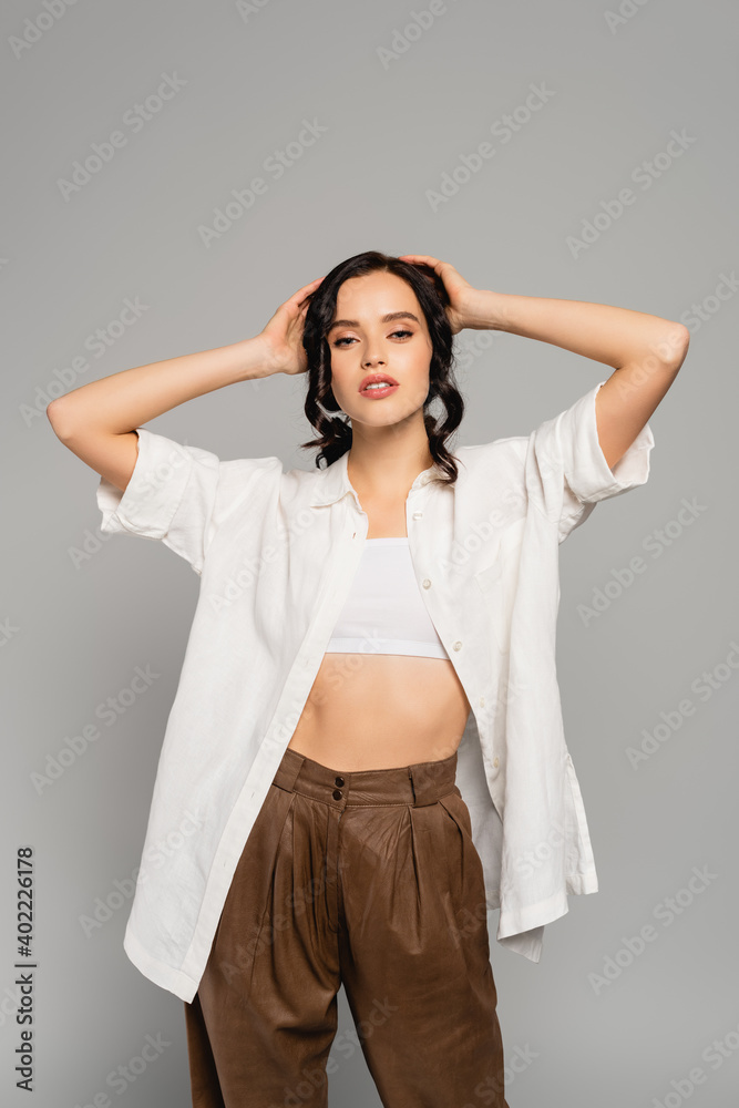 Front view of woman in clothes looking at camera while holding hands near head isolated on grey Front view of woman in clothes looking at camera while holding hands near head isolated on grey