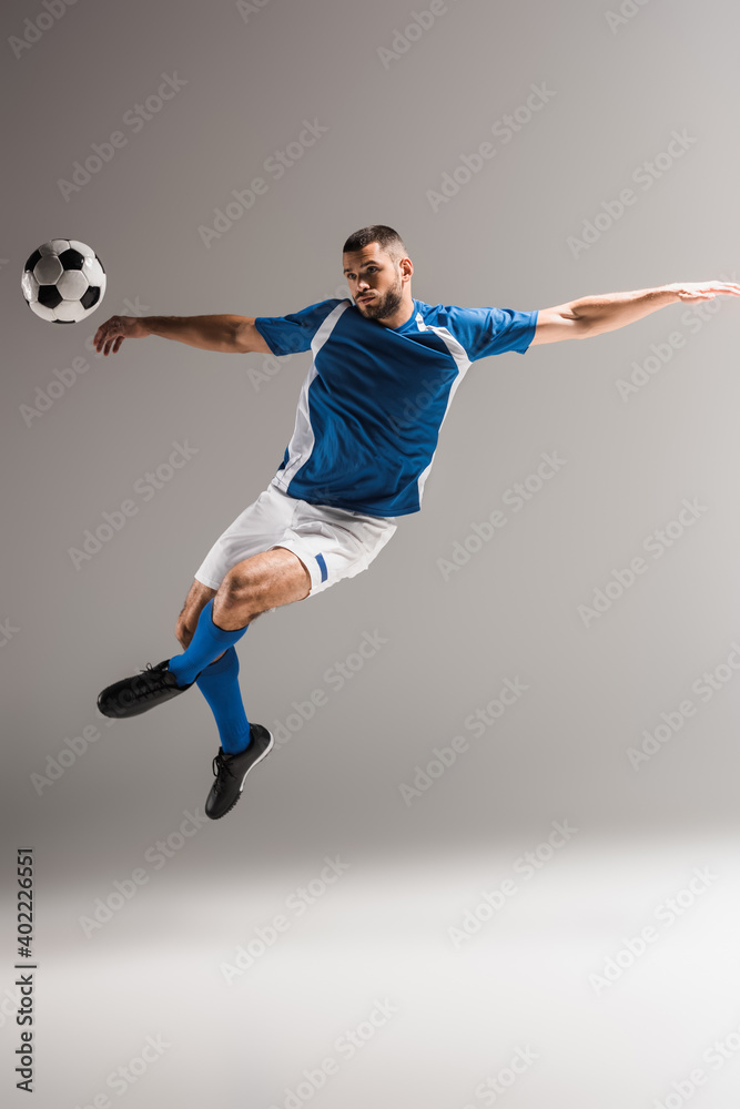 Athletic man jumping with crossed legs near football on grey background