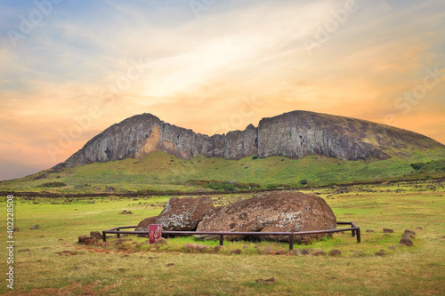 Fallen Moai statue at the Ahu Tongariki ceremonial center on Easter Island, covered by a colorful sunset sky, against the crater of the Rano Raraku volcano. photo