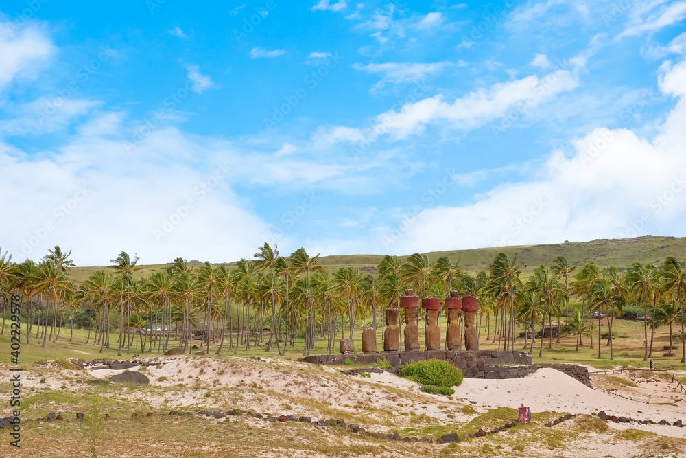 Panoramic view of the palm grove at the Anakena beach and Moai statues at the Ahu Nao-Nao ceremonial center on Easter Island, against a blue sky.