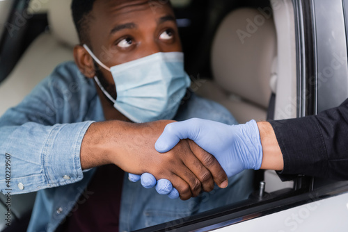Police officer in latex glove handshaking with african american driver in medical mask on blurred background in car.