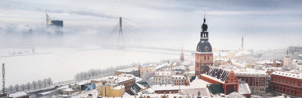Panoramic aerial view of the Riga old town and Daugava river from St. Peter's Church in winter. Morning fog, snow-covered cable-stayed bridge, cathedral, houses. Latvia. Landmarks, sightseeing, travel - obrazy, fototapety, plakaty 
